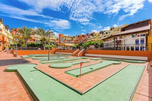 Ein Minigolfplatz mit grünen und orangefarbenen Spielbahnen, umgeben von mehrstöckigen Gebäuden mit Balkonen und Palmen.