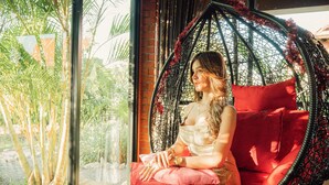 A person sitting in a hanging chair with red cushions, positioned near a window with a view of palm trees and a brick wall.