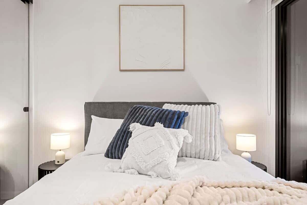 Een net opgemaakt bed met een witte deken en meerdere kussens, twee nachtlampjes en een ingelijst beeld boven het bed.