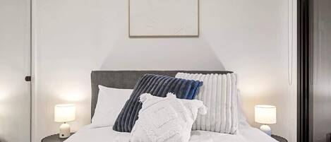 Een net opgemaakt bed met een witte deken en meerdere kussens, twee nachtlampjes en een ingelijst beeld boven het bed.