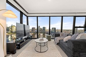 A modern living room with a large TV, comfortable seating, and a view of a city skyline through large windows. - Waterfront 2BR Marina Tower, Rooftop Pool+Parking (Docklands)