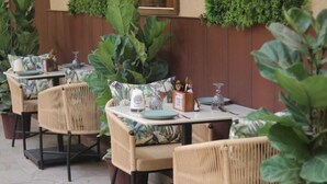 An outdoor dining area with wicker chairs and tables, surrounded by large potted plants. - Lush Premier Hotel (Kanpur)
