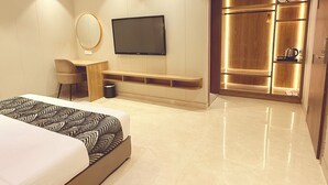 A modern hotel room with a large bed, a flat-screen TV mounted on the wall, a seating area with a small table and chair, and a walk-in closet with illuminated shelves. - Lush Premier Hotel (Kanpur)