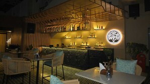 An outdoor bar area with wicker furniture, a bar counter with bottles, and a decorative wall with a circular emblem. - Lush Premier Hotel (Kanpur)