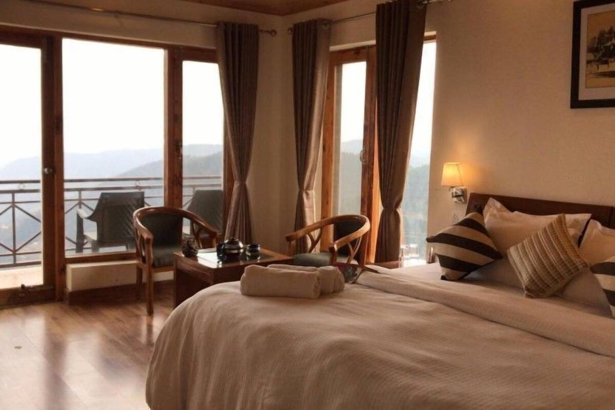 Une chambre d’hôtel avec un grand lit, deux fauteuils, une petite table et une vue sur les montagnes grâce à de grandes fenêtres.