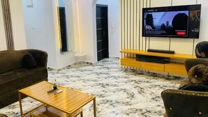 A modern living room with a flat-screen TV mounted on the wall, a wooden coffee table, a sofa, and a TV stand with a TV. - Lakefront 2-BR-Apt With 24Hrs Power + Starlink Wi-Fi (Lagos)