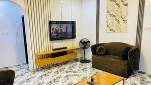 A modern living room with a flat-screen TV mounted on a wall, a brown sofa, a wooden coffee table, and a marble-patterned floor. - Lakefront 2-BR-Apt With 24Hrs Power + Starlink Wi-Fi (Lagos)