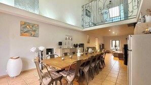 A spacious dining area with a long wooden table and cushioned chairs, a kitchen in the background, and a decorative railing above. - Atypical enitre house with character, large volumes, tastefully decorated (Bourdons-sur-Rognon)