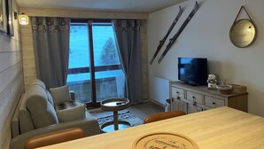 A cozy room with a sofa, a dining table, a TV, and a view of the outdoors through a window. - Le corbier Fully renovated 4-5 person apartment, at the foot of the slopes, view. (Villarembert)