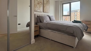 Een moderne slaapkamer met een groot bed, een nachtkastje, een dressoir en een uitzicht op gebouwen door een groot raam.