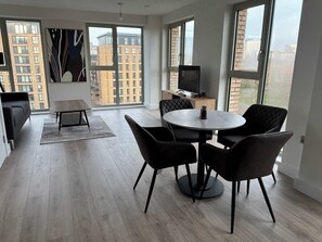Ein modernes Wohnzimmer mit einem runden Esstisch, schwarzen Stühlen, einer Couch und einem Fernseher auf einem Schrank. Große Fenster bieten Blick auf Gebäude im Freien.