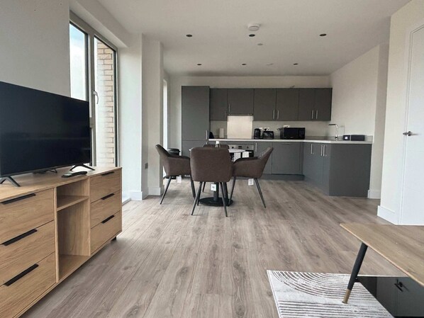 Eine moderne Küche mit grauen Schränken, ein Essbereich mit Tisch und Stühlen sowie ein Fernseher auf einem Holzboden.