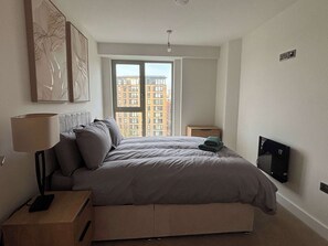 Ein Schlafzimmer mit einem Bett, einem Nachttisch, einer Lampe, einem Fernseher und einem Fenster mit Blick auf die Stadt.