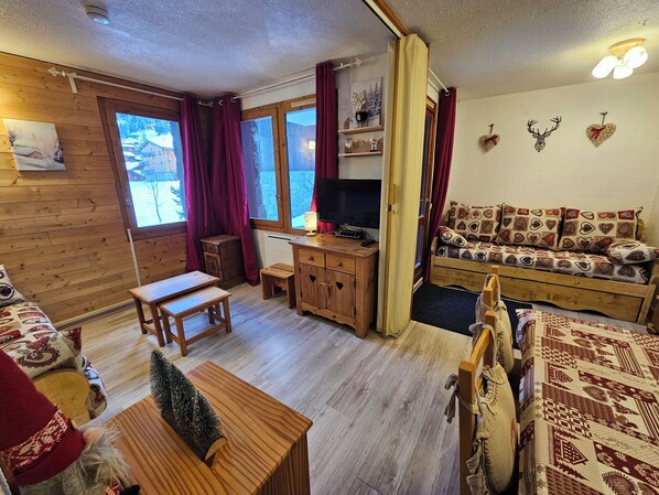Ein gemütliches Blockhaus-Interieur mit Holzboden, einer Couch mit gemusterten Kissen, einem kleinen Holztisch, einem Fernseher und einem Fenster mit Blick auf eine verschneite Landschaft.