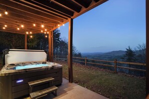 A hot tub under a wooden pergola with string lights, overlooking a fenced yard and distant mountains. - Dog-friendly Retreat with Outdoor Sauna & Hot Tub. Golf and hiking trails nearby (Blue Ridge)