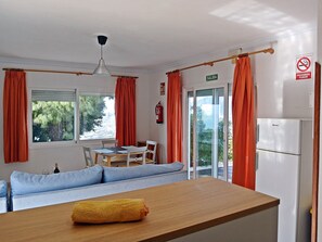 Ein Zimmer mit Holztisch, einer Couch, einem Esstisch mit Stühlen, einem Kühlschrank und Blick auf Grünflächen.