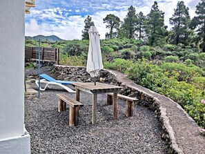 An outdoor seating area with a table and benches, surrounded by a rocky garden and trees. - Een knus vakantiehuis met jacuzzi en prachtig uitzicht op de Atlantische oceaan. (El Paso)