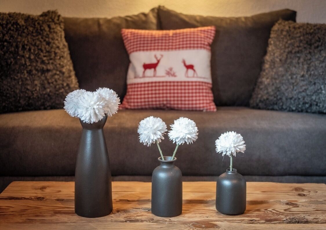 Tre grå vaser med hvide blomster på et træbord foran en brun sofa med en rød og hvid ternet pude.