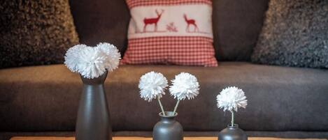 Tre grå vaser med hvide blomster på et træbord foran en brun sofa med en rød og hvid ternet pude.