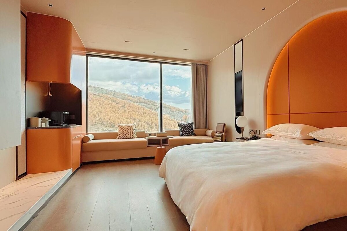 A modern hotel room with a large bed, a sofa, a desk, and a view of the mountains.