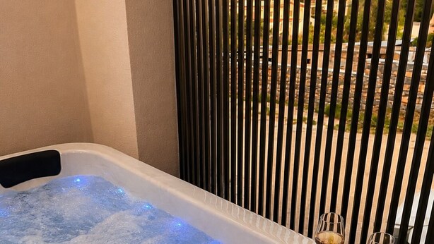 Un bain à remous avec deux verres de vin, un panneau de commande et une vue à travers des stores verticaux.