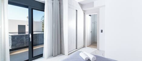 Une chambre d’hôtel moderne dotée d’une grande porte-fenêtre coulissante donnant sur un balcon, d’un dressing et d’une salle de bain avec douche et toilettes.
