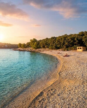 Une plage de galets bordée d’une eau turquoise et limpide, une petite cabane en bois et, en arrière-plan, une colline boisée.