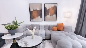 Una sala moderna con un sofá seccional gris, dos mesas redondas con bases negras y doradas, una planta en maceta y arte abstracto en la pared.