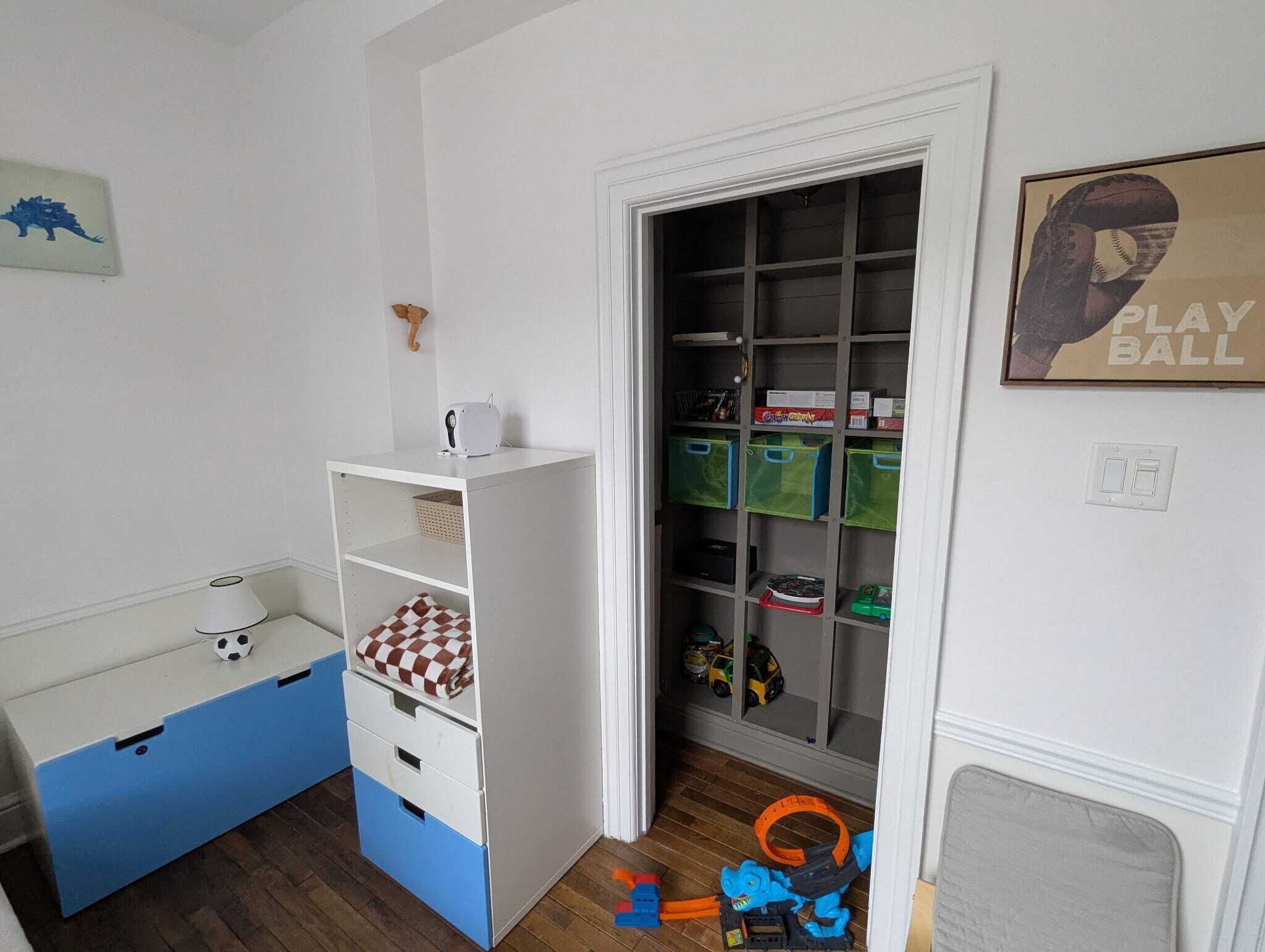 Un cuarto de juegos infantil con un escritorio blanco y azul, un mueble para guardar juguetes y una puerta que da a una zona de juegos.