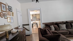 A living room with a brown sofa, a coffee table, and a lamp. There is a door leading to a kitchen and a dining area.