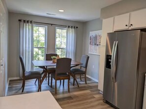 A kitchen with a dining table, chairs, a refrigerator, and a window with curtains.