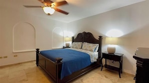 A bedroom with a bed, a ceiling fan, two bedside lamps, and a nightstand. - Rainbow Rise: Oceanfront, Sunsets, Secluded beach, Backup Generator! (Cupecoy)
