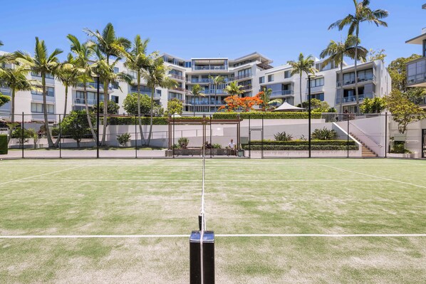 A tennis court with a net in the foreground, surrounded by a fence. In the background, there is a multi-story building with balconies and palm trees. - Solstice by the Sea (Burleigh Heads)