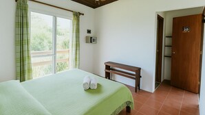 Ein Zimmer mit einem Bett, einer Bank, einem Deckenventilator, einem Fenster mit Vorhängen und einer Tür mit einer Nummer.