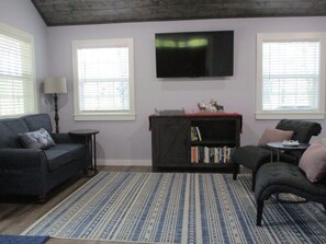 A living room with a sofa, a coffee table, a bookshelf, and a TV mounted on the wall. - The Americana - A Modern & Rustic Cabin with a Woodsy View (Andrews)