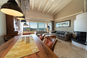 Un vaste espace de vie comprenant une grande table à manger en bois, des fauteuils en cuir, une cheminée et une vue sur les montagnes.