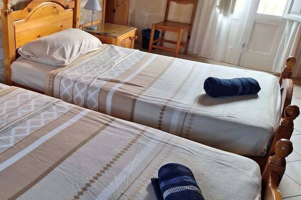 Two wooden beds with patterned bedding, a wooden nightstand, and a chair in a room with a stone wall.