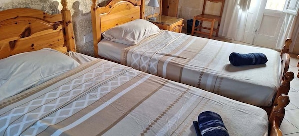 Deux lits en bois avec une literie à motifs, une table de chevet en bois et une chaise dans une pièce dont un des murs est en pierre.