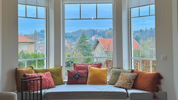 A room with a built-in bench and three large windows offering a view of a town with red-roofed houses and trees. - Villa Mifa 5 minutes from the center with Lagoon Pool (Sarlat-la-Canéda)