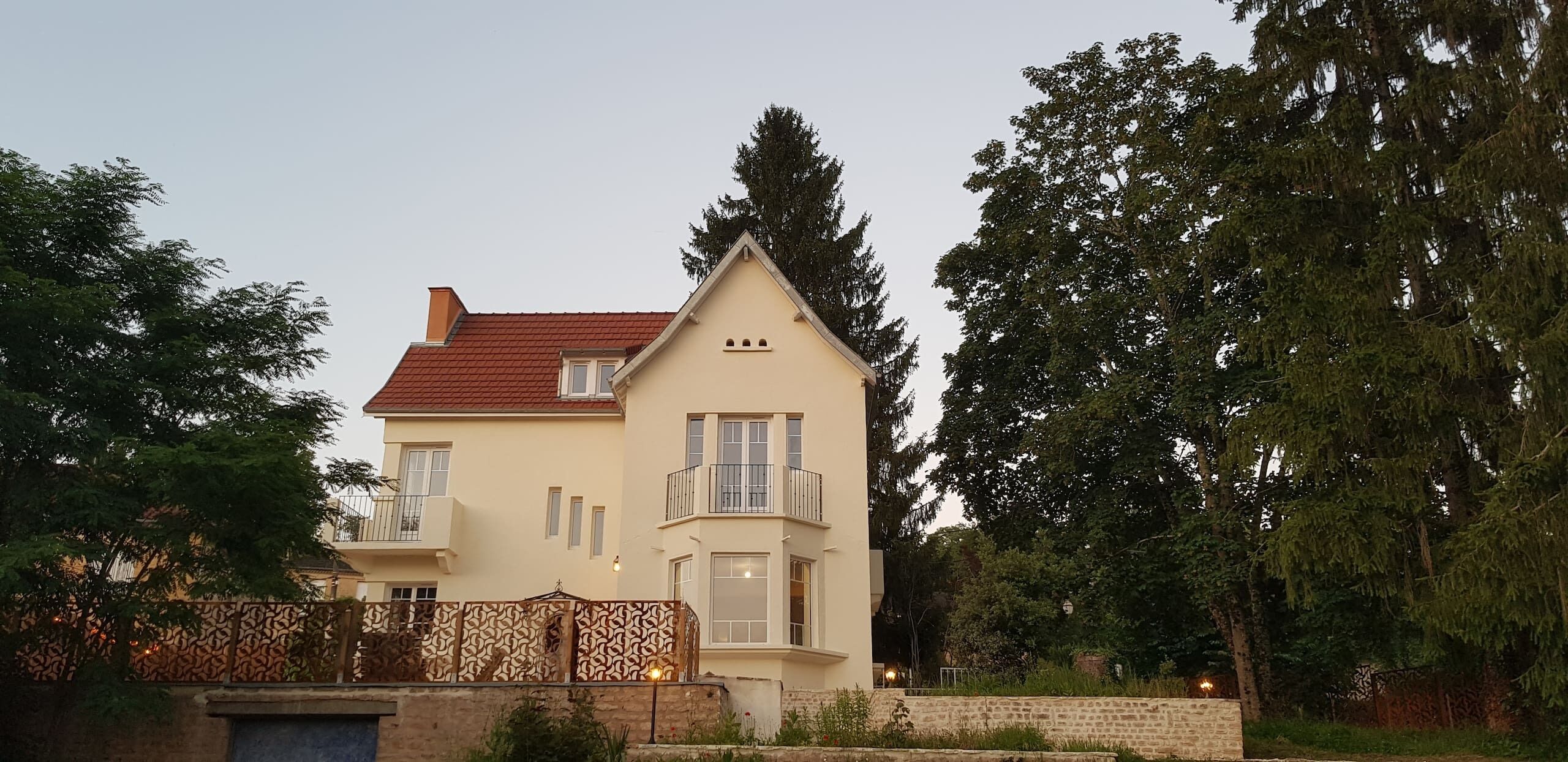 A two-story house with a red roof, a balcony on the second floor, and a stone wall in front.
