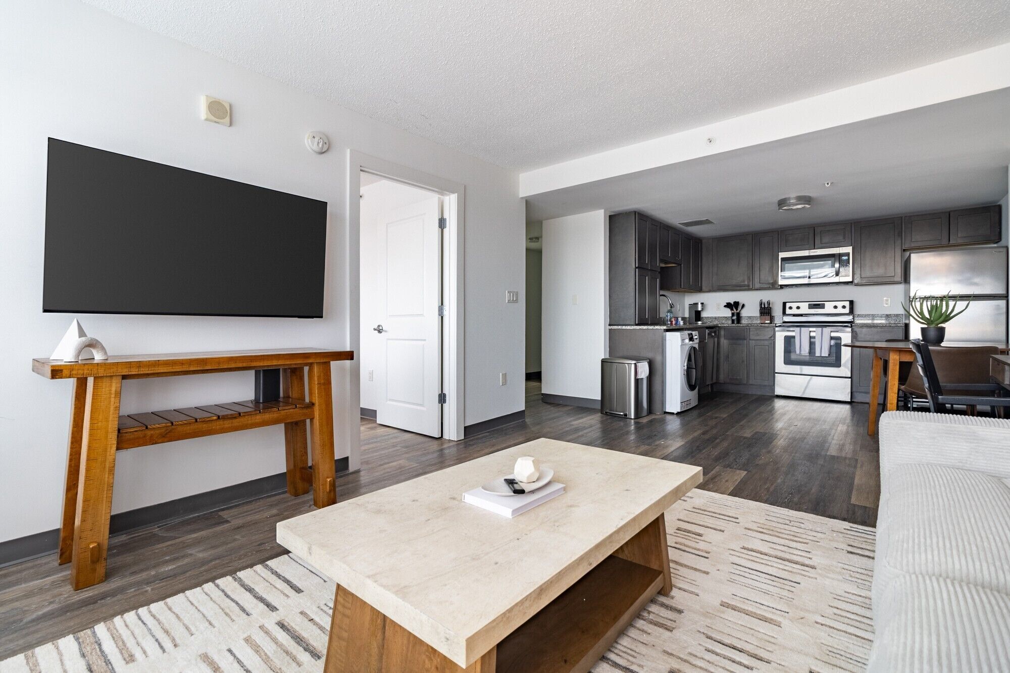 A modern living room with a flat-screen TV, wooden console table, and a beige coffee table. A kitchen with dark cabinets and stainless steel appliances is visible in the background.