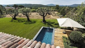 Una piscina con cubierta de madera, rodeada de césped cuidado y olivos.