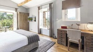 Un dormitorio con cama, escritorio, silla, ventana y vistas a la vegetación exterior.