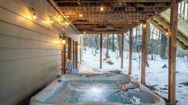 Un jacuzzi bajo una pérgola de madera con luces colgantes, rodeado por un paisaje nevado.