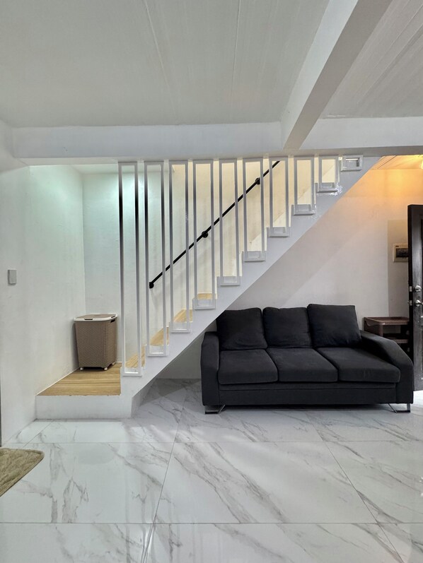 A modern living room with a black sofa, a staircase with white railings, and a marble floor.