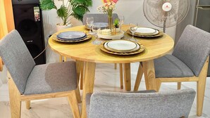 A dining area with a round wooden table set for four, grey upholstered chairs, a large flat-screen TV mounted on the wall, a standing fan, and a potted plant.