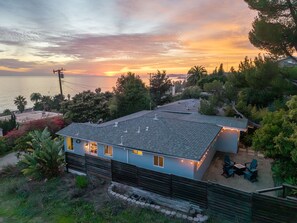Ein Wohnhaus mit geneigtem Dach, einer Terrasse mit Sitzgelegenheiten und Blick auf das Meer bei Sonnenuntergang.