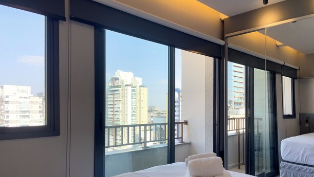 Een hotelkamer met een groot bed, uitzicht op hoge gebouwen en een balkon met een hekwerk.