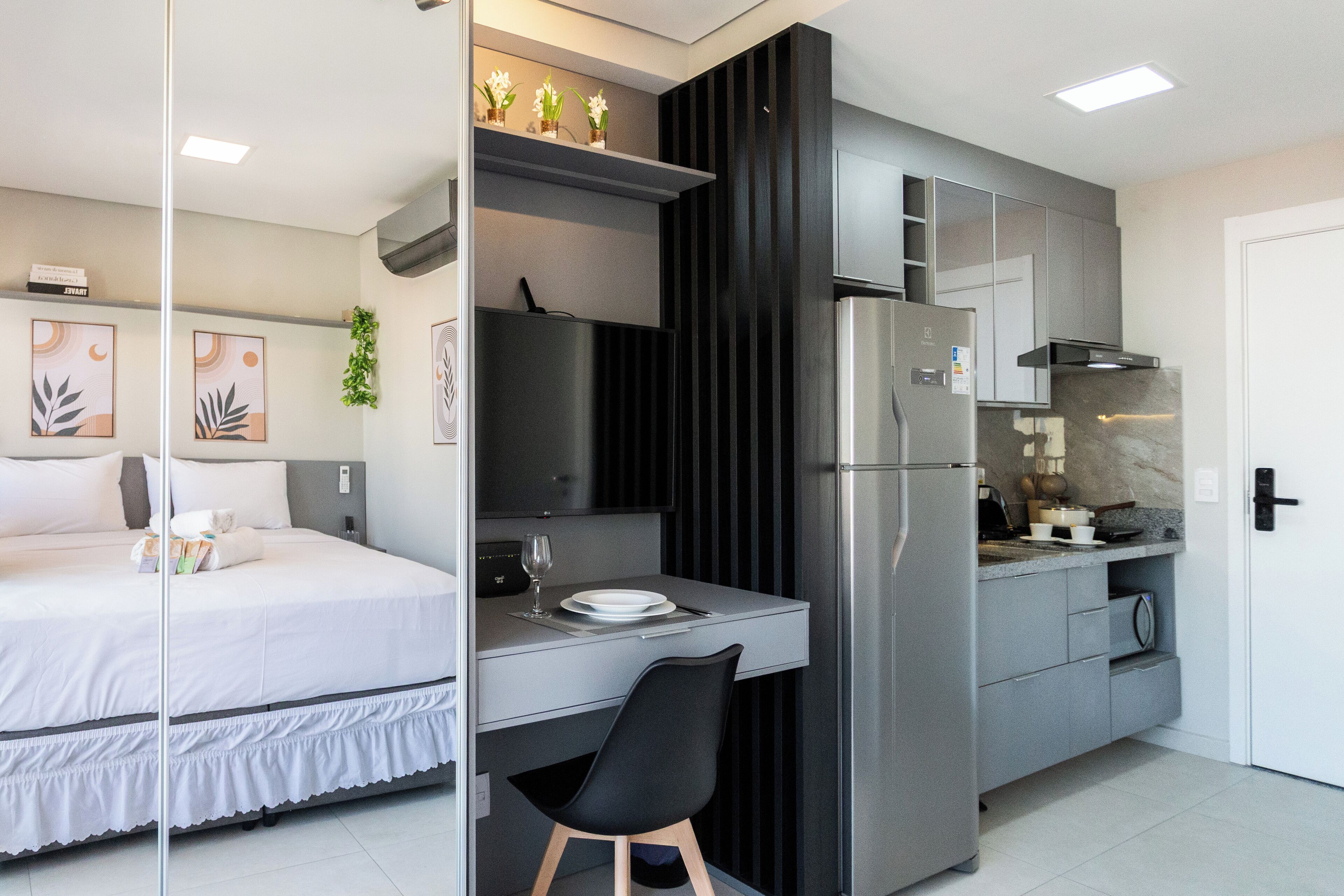 Una habitación de hotel moderna con una cama, una pequeña mesa de comedor con una silla, una kitchenette con un refrigerador y una puerta blanca.