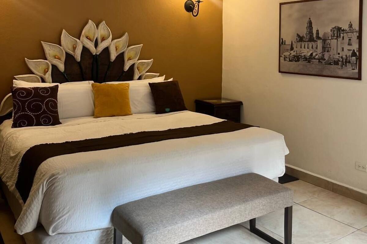 Ett hotellrum med en säng, en bänk, en vägglampa och en inramad tavla.